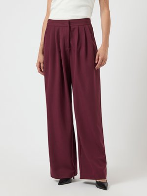 Burgundy vida byxor med veck, tillverkade av slät tyg. Har en hög midja design, visad ihop med svarta spetsiga klackar.
