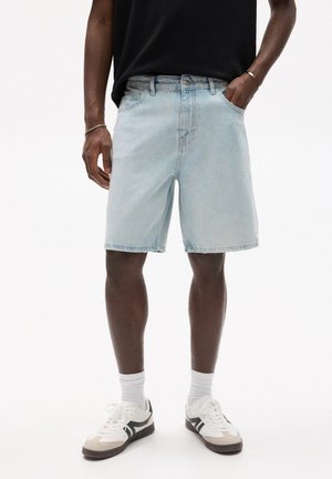 Mann trägt hellblaue Jeansshorts, schwarzes Shirt, weiße Socken und weiße Sneaker mit schwarzen Details und steht mit einer Hand in der Tasche.