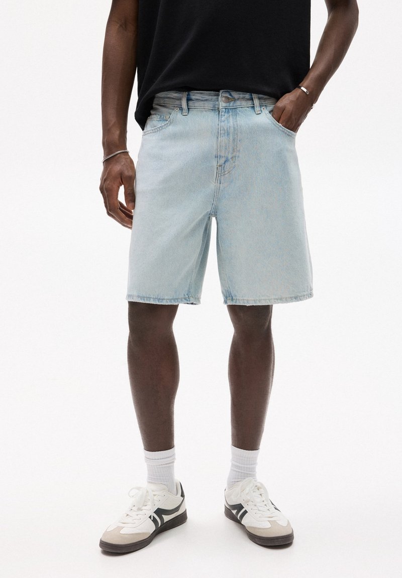 Homme portant un short en jean bleu clair, une chemise noire, des chaussettes blanches et des baskets blanches avec des détails noirs, debout avec une main dans la poche.