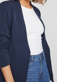 Cardigan bleu marine à ouverture devant et manches trois-quarts; associé à un top blanc ajusté et un jean taille haute. Texture de tissu douce.