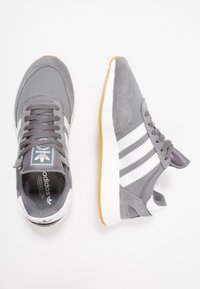 Grå sneakers med vita ränder och en texturerad ovansida. Utrustad med en gummisula och en vadderad tunga. Material av mocka och meshtyg.