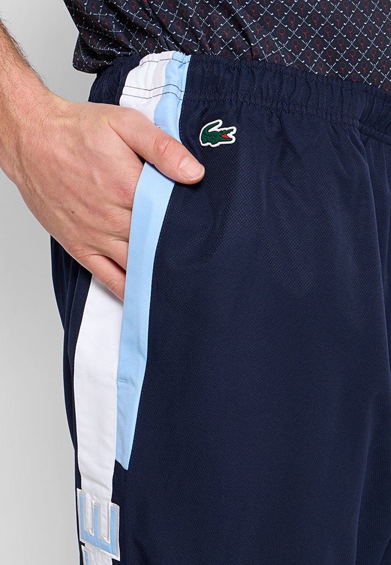 Shorts de sport marine avec des bandes latérales bleu clair et blanches, une taille élastique et un logo Lacoste vert sur la poche gauche.