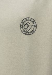 Jasnoszara bawełniana koszulka z okrągłym czarnym logo zawierającym napisy "Club Jack & Jones" oraz "Since 1990". Gładka tekstura, design z okrągłym dekoltem.