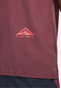 Camiseta de senderismo Nike en color burdeos, con una tela texturizada, un logo triangular en rojo y un dobladillo con aberturas laterales para mayor comodidad en el movimiento.