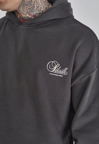 Felpa con capuccio grigio carbone realizzata in tessuto morbido, con un logo bianco sul petto. Questo design include un cappuccio con coulisse e una vestibilità comoda.