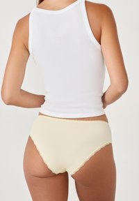 Bragas de algodón amarillo claro con encaje, combinadas con una camiseta blanca ajustada, mostrando la vista posterior y una textura suave.