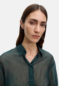 Ciemnozielona, przezroczysta koszula z kołnierzykiem zapinanym na guziki, z małymi zielonymi ozdobami i miękką teksturą.