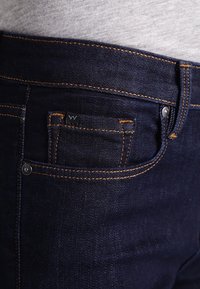 Mörkblå denimjeans med orange sömmar, en liten framficka och en metallknappdetalj vid midjan.