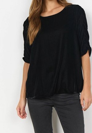 Femme portant un haut en velours noir à manches larges froncées et un pantalon ajusté gris foncé sur un fond blanc uni.