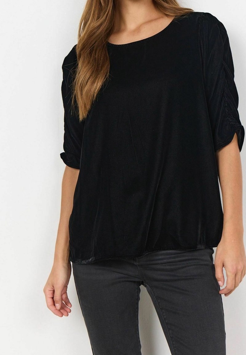 Femme portant un haut en velours noir à manches larges froncées et un pantalon ajusté gris foncé sur un fond blanc uni.