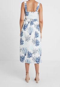 Femme portant une robe midi blanche avec des motifs de feuilles bleues, neckline carré au dos, et des sandales à talons beiges, se tenant devant un fond uni.