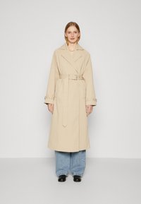 Beige trench coat med belte i livet, brede klaffeskjørt og mansjettstropper, båret over løse denimjeans og svarte sko. Glatt stoff og skreddersydd passform.