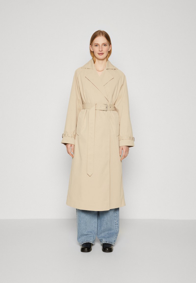 Beige trench coat med belte i livet, brede klaffeskjørt og mansjettstropper, båret over løse denimjeans og svarte sko. Glatt stoff og skreddersydd passform.