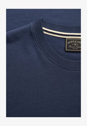 Granatowy t-shirt z okrągłym dekoltem, prążkowanym kołnierzem oraz czarną metką z napisem „Superdry Premium Outfitter New York” wewnątrz kołnierza.