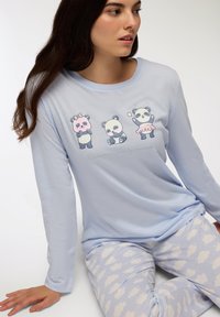 La camicia azzurra a maniche lunghe presenta quattro design di panda cartoon; abbinata a pantaloni azzurri con motivi di nuvole bianche, creando un look accogliente.