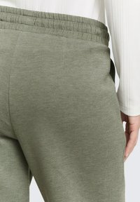 Joggingbroek in olijfgroen met een zachte textuur, elastische tailleband en zijzakken, gedeeltelijk zichtbaar met een witte, geribbelde, lange mouwen top.
