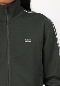 Chaqueta de cremallera verde oscuro con cuello alto, tejido texturizado, franjas laterales blancas y pequeño logo de cocodrilo bordado en el pecho izquierdo.