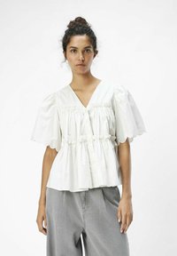 Weiße Baumwollbluse mit V-Ausschnitt, Puffärmeln, gerafftem Bund und Knöpfen vorne. Mit gewelltem Ärmelrand und lockerer Passform.