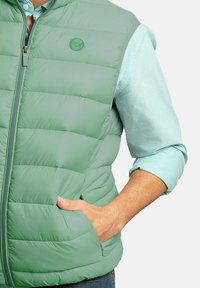 Chaleco acolchado verde claro con cremallera frontal, con costuras horizontales y bolsillos laterales. Llevado sobre una camisa azul clara.