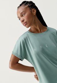 Svetlozelené tričko s krátkym rukávom vyrobené z hladkej tkaniny s rolovanými rukávmi a jemným bielym logom. Mäkka textúra a uvoľnený strih.