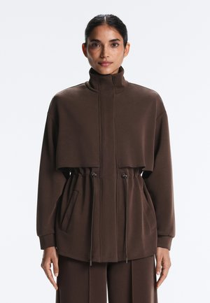 Frau mit zurückgebundenem dunklem Haar, die eine braune Jacke mit Reißverschluss, hohem Kragen und verstellbaren Taillen-Kordeln trägt, steht vor einem einfarbigen Hintergrund.