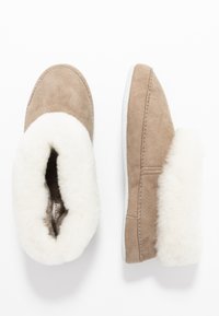 Tan suede slippers with white shearling lining and fold-over cuff, rounded toe, and contrasting white sole. Subtle stitching accents.