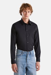 Camicia nera con bottoni e maniche lunghe, caratterizzata da una texture liscia e un colletto classico, abbinata a jeans blu sbiaditi.