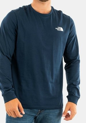 Mann trägt ein dunkelblaues Langarmshirt von The North Face mit Logo auf der Brust, kombiniert mit blauen Jeans, vor einem schlichten Hintergrund.