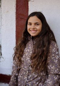 Bruine jas met bloemenpatroon, met een hoge kraag, knopen aan de voorkant en lange mouwen. Het model heeft lang, golvend haar en lacht.