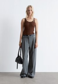 Brown ribbed tank top with buttons, grey pleated trousers, black woven handbag, and black shoes, set against a plain background.