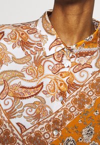 Chemise boutonnée avec un motif complexe de paisley et floral en tons orange, blanc et gris ; tissu lisse avec un col classique.