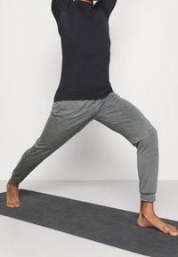 Person in black long-sleeve top and gray jogger pants stretching legs in a yoga pose on a dark gray mat against white background.