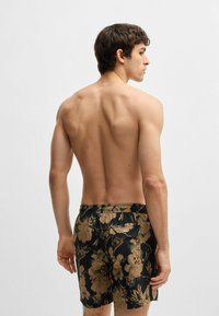 Musta ja kultaiset kukkakuosiset uimaportsit, joissa on joustava vyötärö, takatasku ja sileä, kevyt kangas. Malli näkyy takaa.