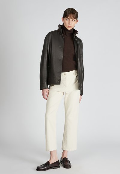Giacca di pelle nera con colletto alto, dolcevita marrone scuro, pantaloni color crema e mocassini marrone scuro, esposti su uno sfondo chiaro.