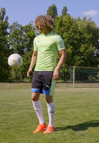 Tricou sportiv cu dungi verzi, pantaloni scurți negri cu detalii albastre, șosete albe și ghete portocalii. O minge de fotbal este așezată lângă persoană.