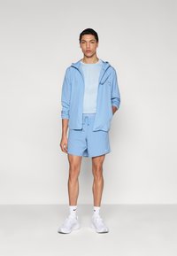 Αθλητική ενδυμασία σε ανοιχτό γαλάζιο χρώμα που περιλαμβάνει ένα ελαφρύ τζάκετ με φερμουάρ και σορτς, συνδυασμένη με λευκά αθλητικά παπούτσια και αντίστοιχες κάλτσες.