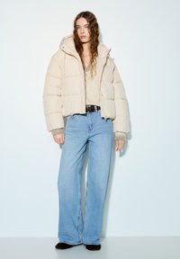 Geacă matlasată bej cu glugă, cu model matlasat, mâneci largi și manșete drepte; combinată cu blugi evazați bleu deschis și un pulover bej.