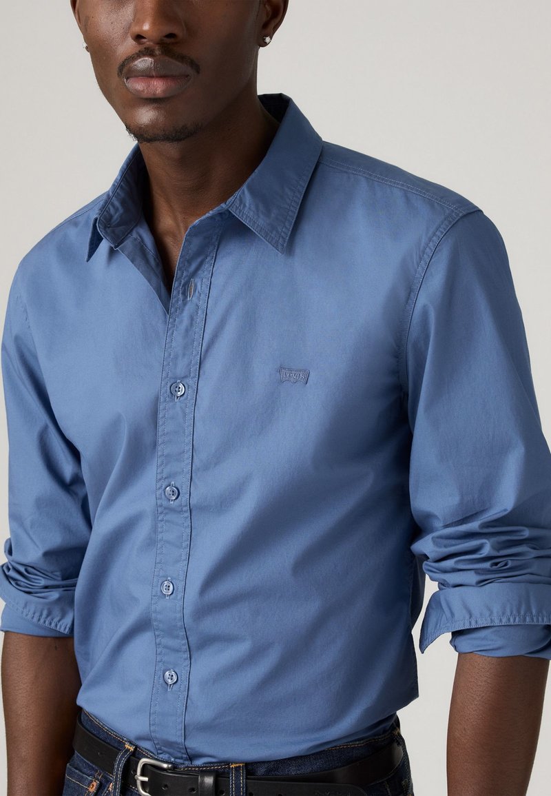 Chemise boutonée bleu clair en coton, avec un col structuré, des manches longues et un petit logo sur la poitrine. Texture lisse.