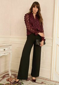 Kvinna med långt brunt hår som bär en genomskinlig bordeauxfärgad blus med prickar, svarta vida byxor, bordeauxfärgade klackar, och håller en svart kuvertväska.