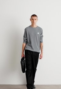 Γκρι μακρυμάνικο πουκάμισο με μικρό λογότυπο, σε συνδυασμό με μαύρα παντελόνια. Το άτομο κρατά μια μαύρη τσάντα, σε έναν μινιμαλιστικό φωτεινό φόντο.