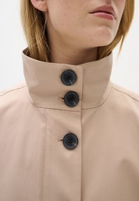 Béžový kabát s vysokým límcem, obsahující tři tmavé knoflíky, jemnou texturu a minimalistický design. Hladká látka s jemnými švy.