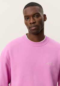 Ροζ φούτερ με στρογγυλή λαιμόκοψη και ριμπ μανσέτες. Διακοσμημένο με κεντημένο κείμενο "Les Deux" σε λευκό χρώμα στην αριστερή πλευρά. Κατασκευασμένο από μαλακό μείγμα βαμβακιού.