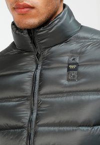 Donsjack in donkergrijs nylon met een hoge kraag en ritssluiting aan de voorkant. Voorzien van een logo-opdruk en doorgestikte voering voor isolatie.