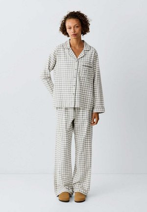 Femme debout portant un haut de pyjama ample à manches longues blanc et gris à carreaux et un pantalon assorti avec des chaussons marron sur un fond uni.