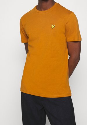 Uomo che indossa una maglietta a maniche corte color senape con un piccolo logo giallo di un uccello sul petto, abbinata a pantaloni scuri, in piedi davanti a uno sfondo semplice.