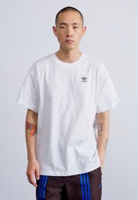Tricou din bumbac alb cu mâneci scurte, având un mic logo negru adidas pe zona stângă a pieptului. Modelul poartă șorturi maro cu dungi albastre.