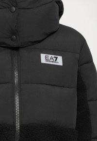 Zwarte pufferjack met capuchon, voorzien van een zachte textuur, een ritsluiting en een logo-opdruk op de borst, afgewerkt met een bontachtige stof.