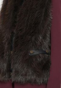 Gilet en fausse fourrure marron foncé avec une finition texturée, comprenant un panneau en cuir noir et des accents en métal doré.