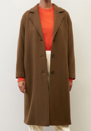 Frau trägt einen langen braunen Wollmantel über einem orangefarbenen Pullover und cremefarbenen Hosen, steht vor einem schlichten hellen Hintergrund.