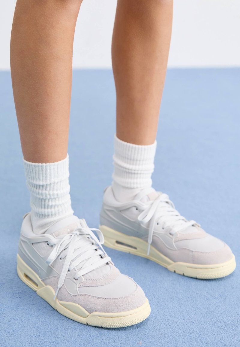 Chaussures de sport gris clair en suède et mesh, lacets blancs, et semelle en caoutchouc jaunie. Portées avec des chaussettes blanches côtelées.